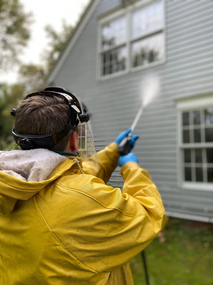 A washer in a yellow rain jacket spraying the upper siding of a house beside a window